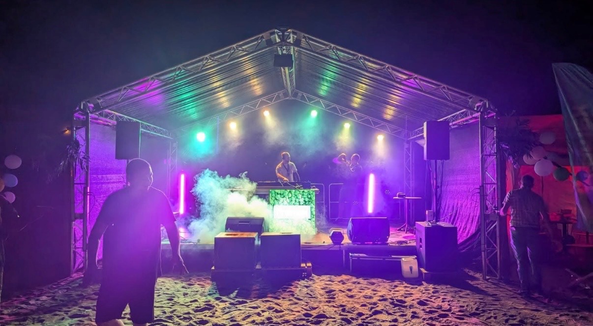 Scena plenerowa z DJ-em i oświetleniem w kolorach zieleni i fioletu, z efektem dymu. Widoczni ludzie na scenie i przed nią, piasek na podłożu.