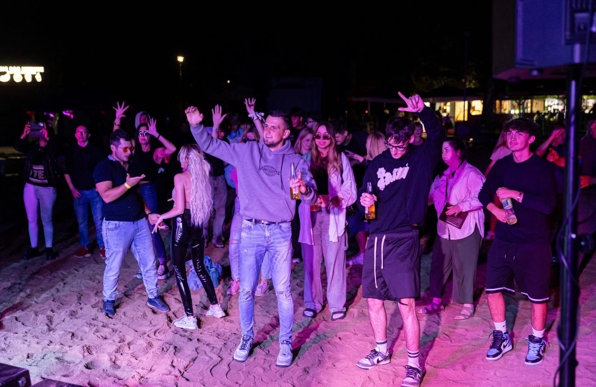 Rozbawiony tłum tańczy na plaży w nocnym oświetleniu, trzymając napoje. Widoczny fragment nagłośnienia. Dynamiczna atmosfera imprezy plenerowej.