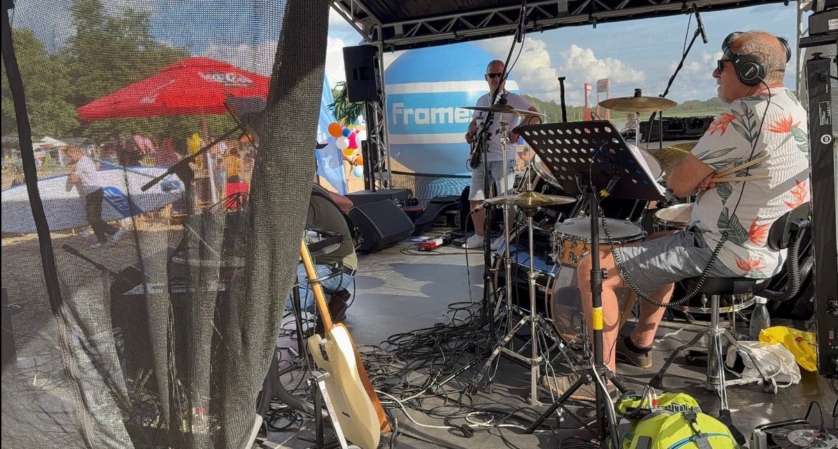 Zespół muzyczny na scenie plenerowej: perkusista w koszuli w kwiaty i saksofonista w tle, widok zza kulis przez siatkę. Scena z nagłośnieniem i instrumentami.