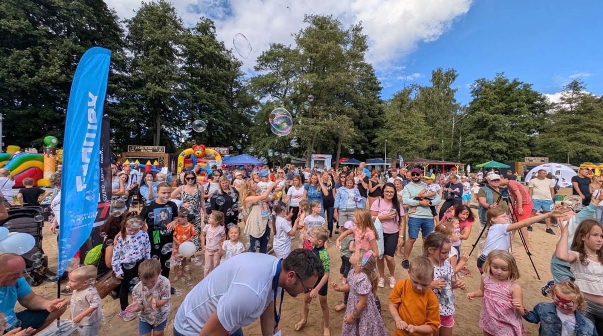 Plenerowa impreza z dmuchańcami, animacjami dla dzieci i stoiskami gastronomicznymi, otoczona zielenią drzew. W tle niebieski baner reklamowy.