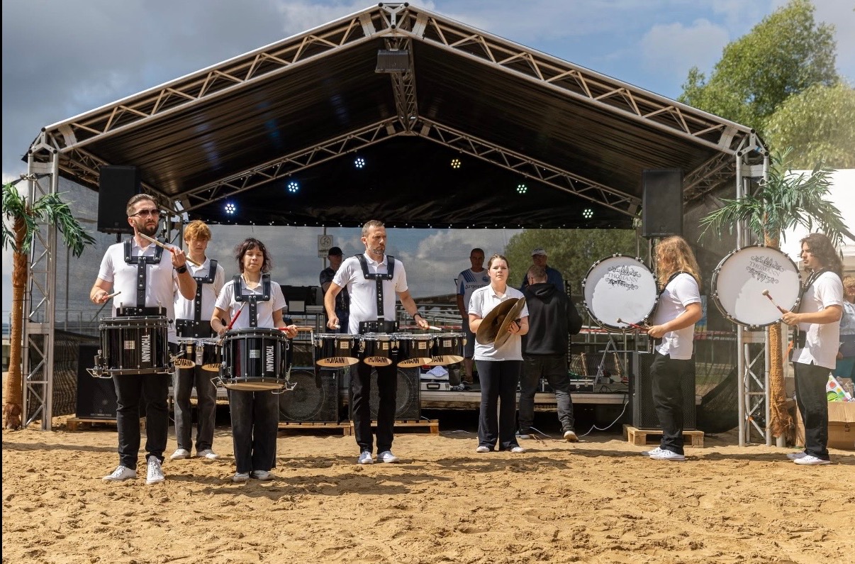 Zespół perkusyjny w białych koszulkach gra na bębnach i talerzach na piaszczystej scenie pod zadaszeniem, z palmami po bokach. Widoczni również widzowie w tle.