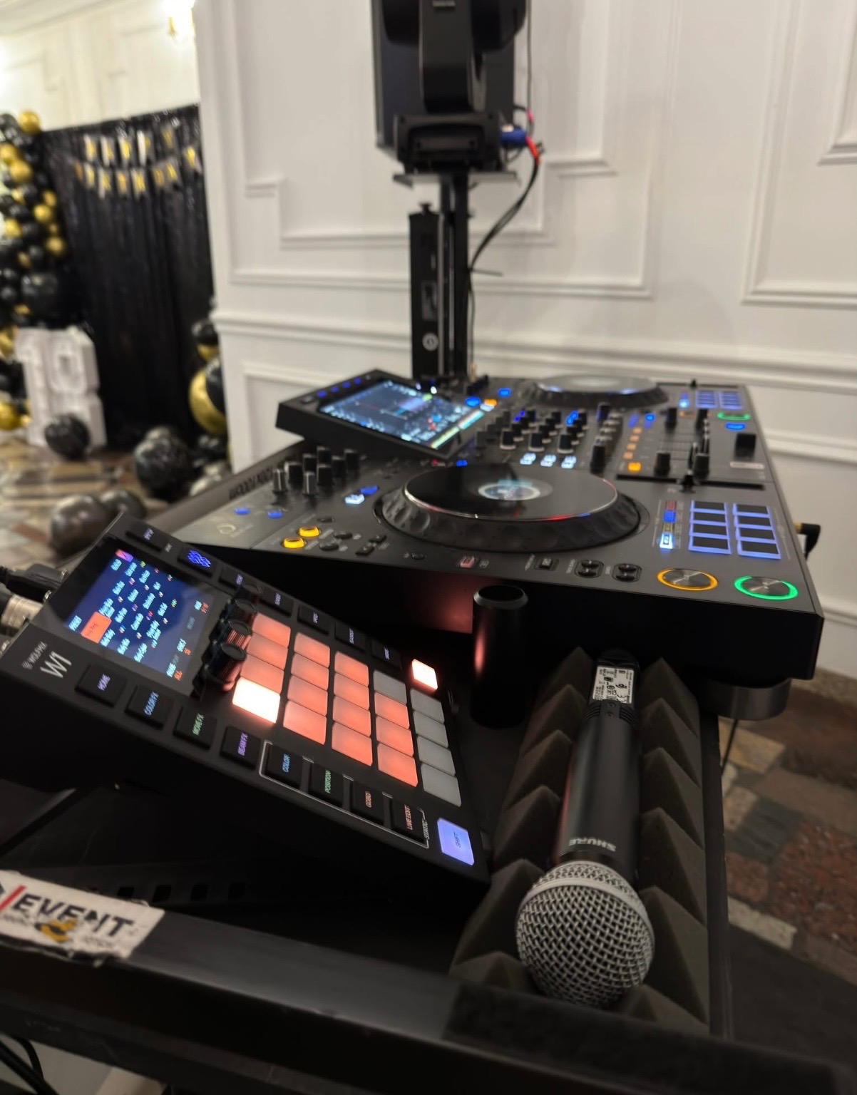 Profesjonalny sprzęt DJ-ski: mikser, kontroler, mikrofon Shure i oświetlenie sceniczne na tle dekoracji imprezowej z czarno-złotymi balonami.