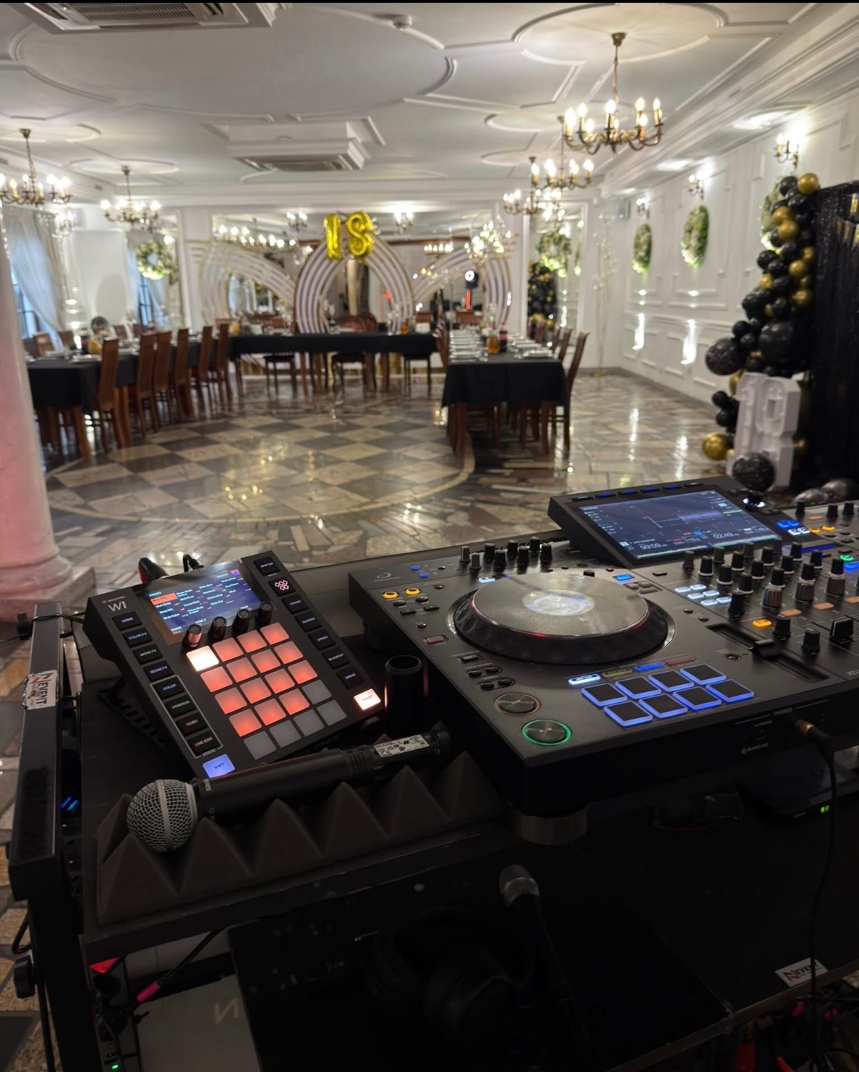 Profesjonalny sprzęt DJ-ski z mikrofonem na tle eleganckiej sali balowej z dekoracjami urodzinowymi, gotowy do obsługi imprezy. Widoczne stoły i krzesła.
