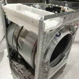 Rozłożona pralka LG z widocznym bębnem i elementami elektronicznymi, prawdopodobnie w trakcie naprawy. Widoczne śruby na podłodze.