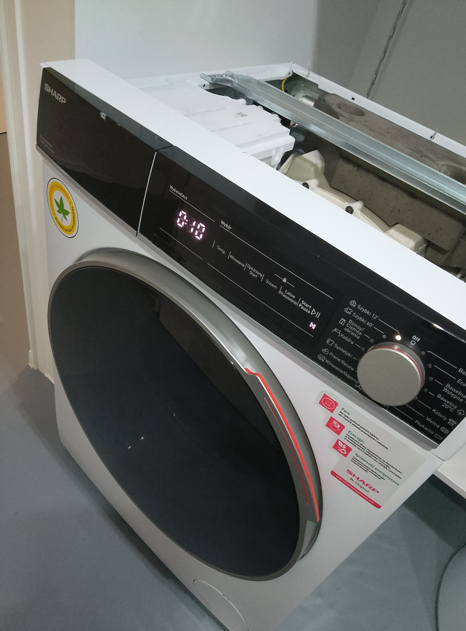 Otwarta pralka Sharp z widocznymi elementami wewnętrznymi, ustawiona pod kątem. Panel sterowania z wyświetlaczem i pokrętłem. Widoczne naklejki informacyjne na obudowie.
