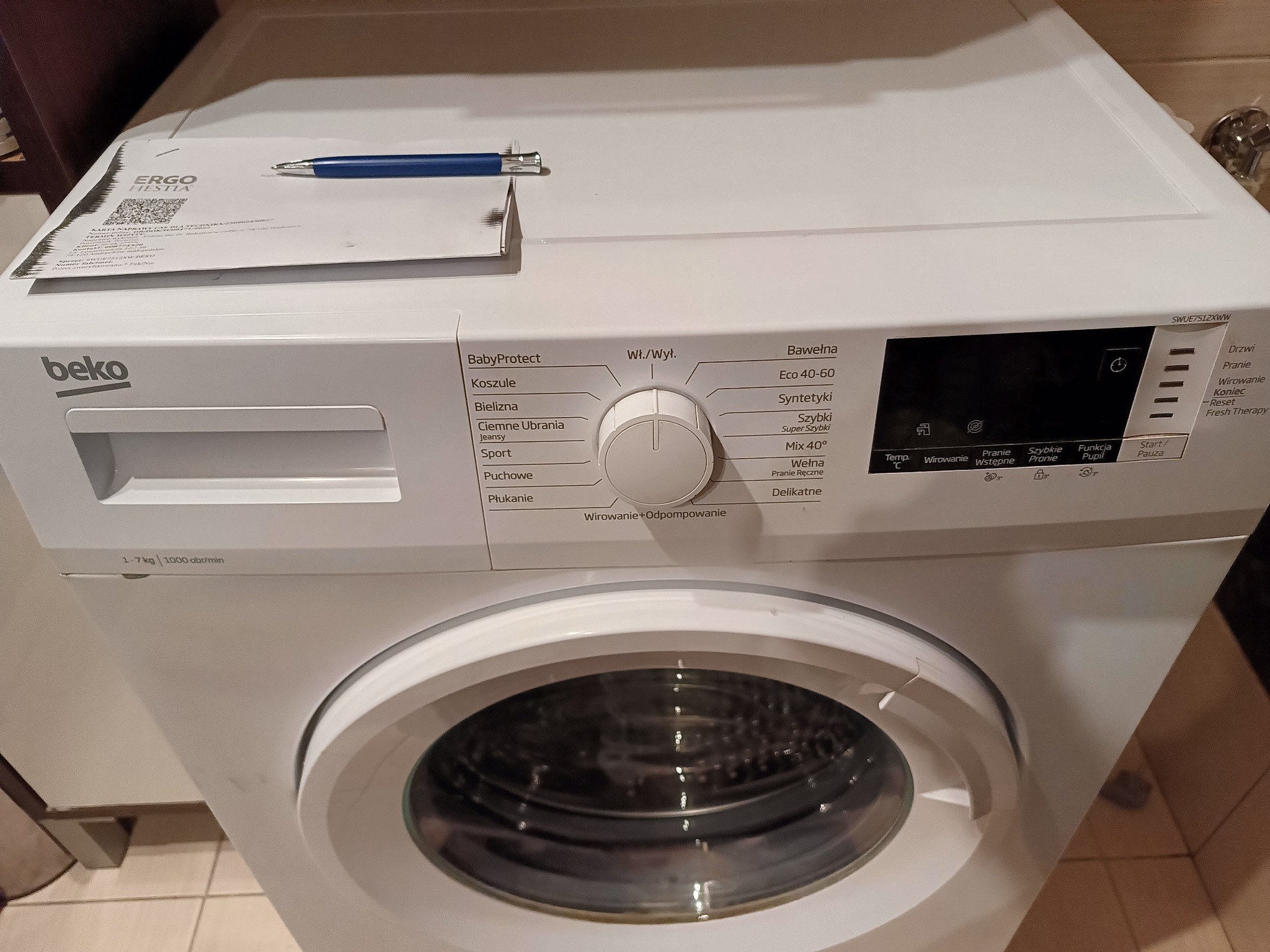 Biała pralka Beko z programami, na niej karta naprawcza Ergo Hestia i niebieski długopis. Widoczny panel sterowania i bęben pralki. Ustawienia prania.