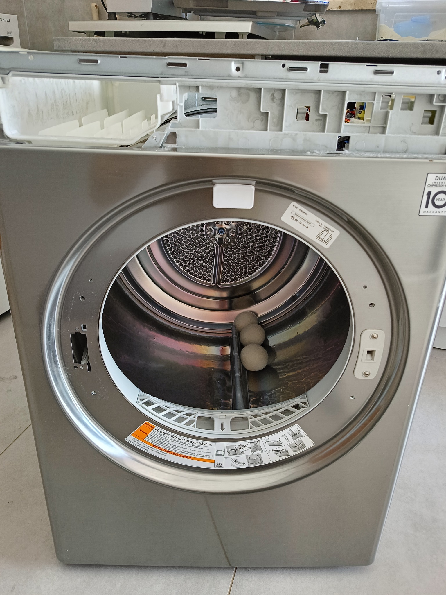 Otwarta suszarka bębnowa LG w kolorze srebrnym, z widocznym wnętrzem bębna i trzema kulami do suszenia. Widoczne instrukcje użytkowania na obudowie.