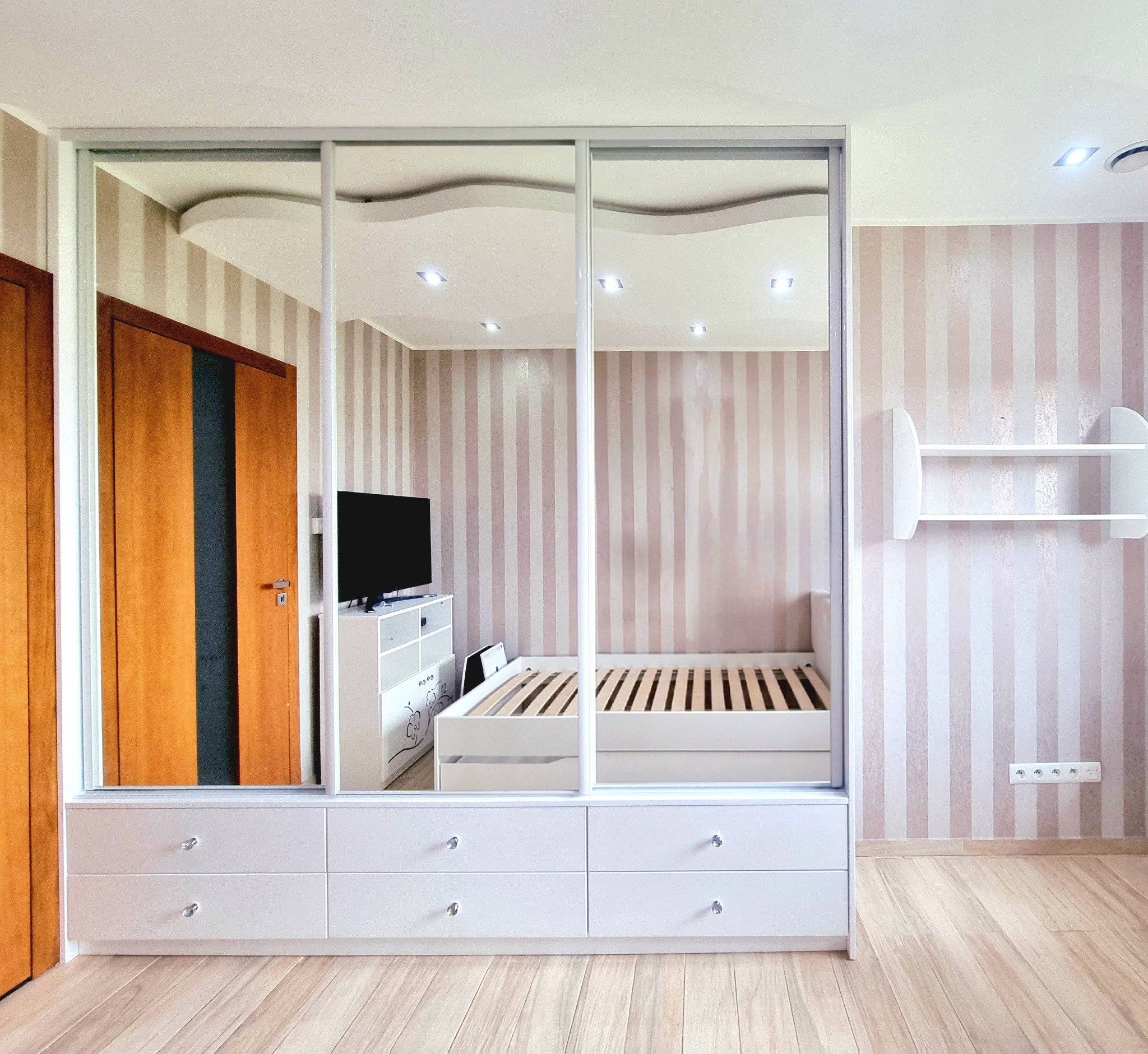 Nowoczesna szafa przesuwna z lustrami w pokoju z łóżkiem i komodą, odbicie wnętrza w lustrzanych drzwiach, paski na ścianie i jasna podłoga.