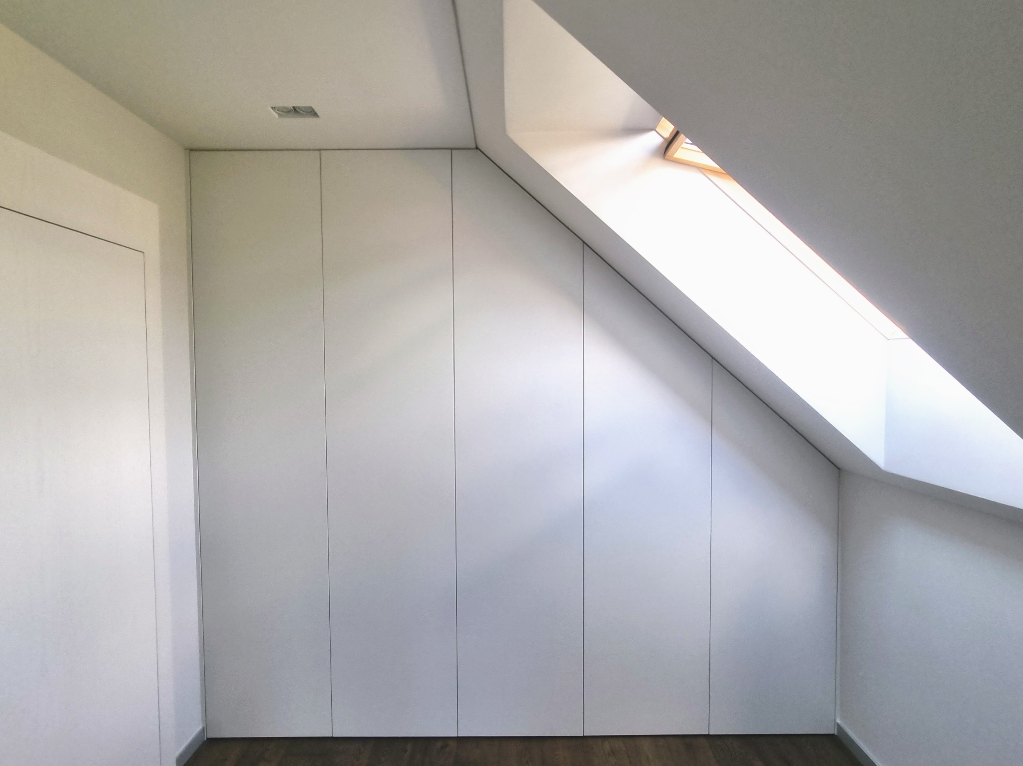 Biała szafa wnękowa pod skosem dachu, idealnie dopasowana do przestrzeni poddasza. Widoczne drzwi bez uchwytów, drewniana podłoga i okno dachowe wpuszczające światło.