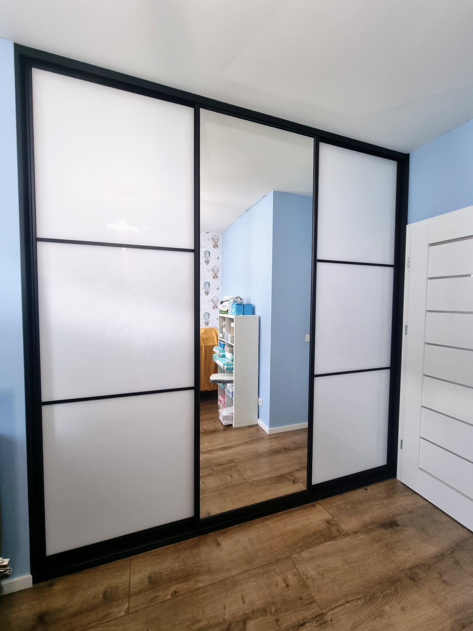 Szafa przesuwna z czarnymi ramami i białymi panelami, jedna część lustrzana, na drewnianej podłodze. Widoczne wnętrze pokoju z niebieskimi ścianami i meblami.