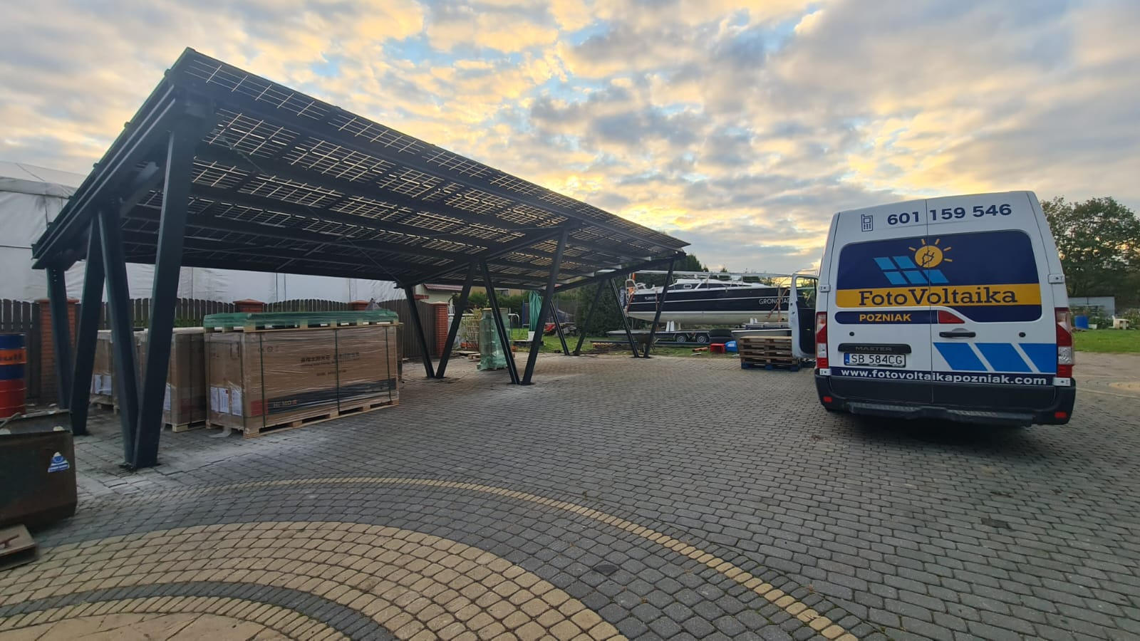 Zadaszenie z panelami słonecznymi, zapakowane panele na paletach i furgonetka z logo firmy. Montaż paneli fotowoltaicznych na parkingu.