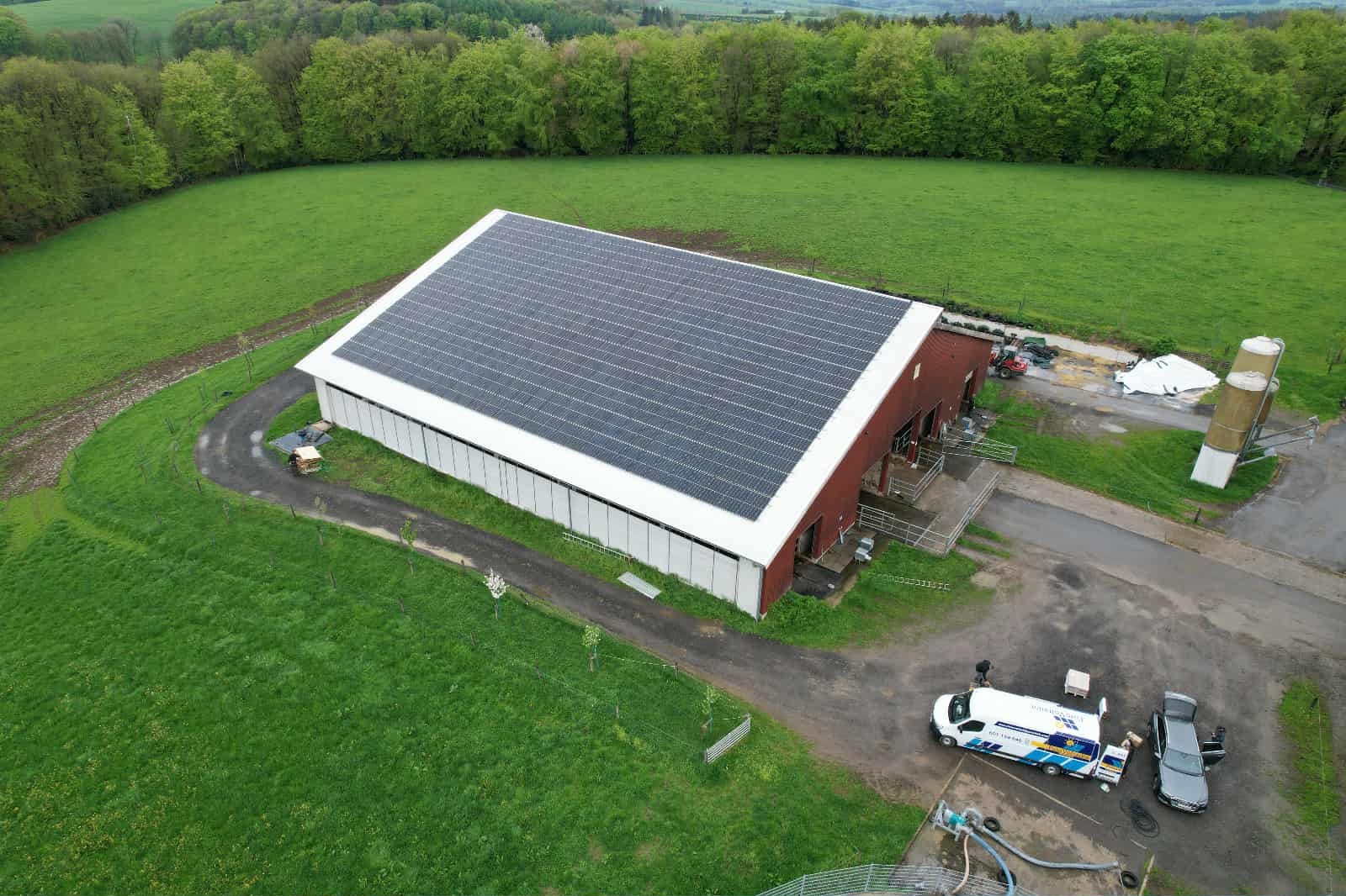 Widok z drona na budynek gospodarczy z panelami słonecznymi na dachu, zaparkowany bus serwisowy firmy i zielone otoczenie.