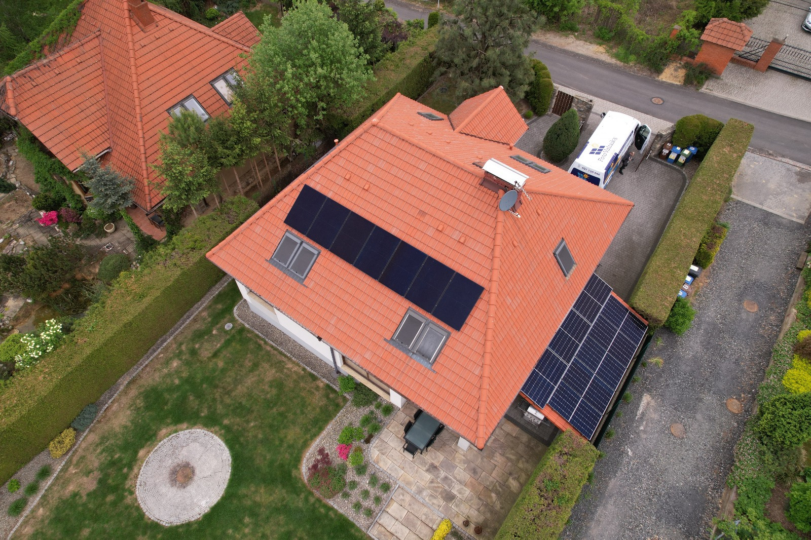 Widok z góry na dom z pomarańczową dachówką i panelami fotowoltaicznymi. Obok domu zaparkowany biały samochód dostawczy z logo firmy.