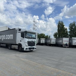 HoldCargo sp. z o.o. - Ciężarówka HoldCargo.com na placu z zaparkowanymi naczepami. W tle błękitne niebo z chmurami i słup wysokiego napięcia. Widok z dołu, perspektywa szerokokątna.