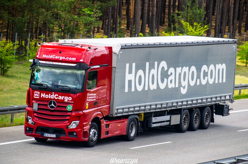 Czerwona ciężarówka marki Mercedes z logo HoldCargo.com na szarej naczepie, jadąca autostradą na tle lasu. Widoczne detale: tablica rejestracyjna, oznaczenia na kabinie.