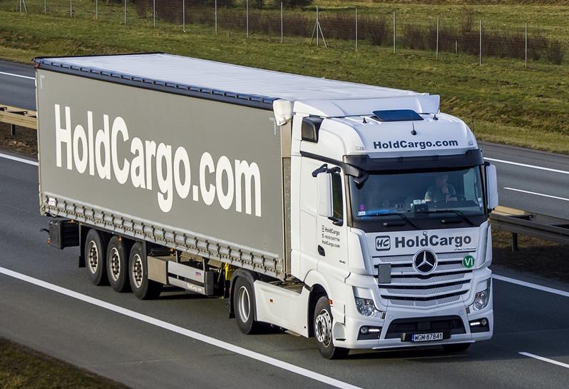 Biała ciężarówka Mercedes z szarą naczepą HoldCargo.com na autostradzie, widok z boku, w tle zielone pola. Rejestracja WGM 87841.