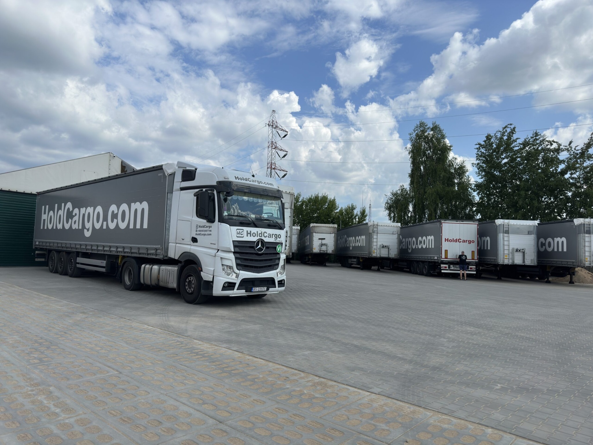 Ciężarówka HoldCargo.com na placu z zaparkowanymi naczepami. W tle błękitne niebo z chmurami i słup wysokiego napięcia. Widok z dołu, perspektywa szerokokątna.