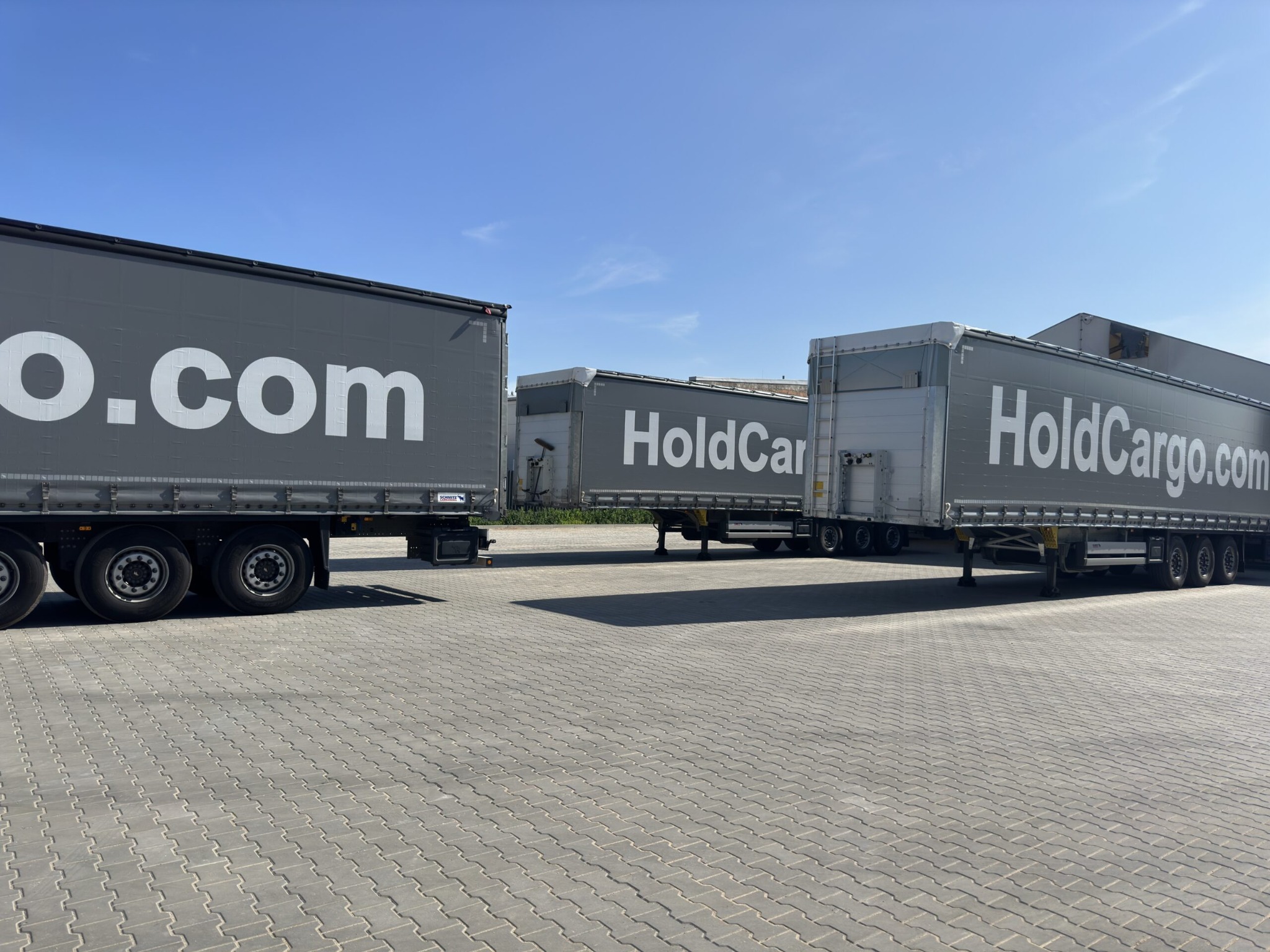 Szare naczepy z logo HoldCargo.com ustawione rzędem na parkingu z kostki brukowej pod błękitnym niebem. Widoczny fragment naczepy z napisem '.com'.