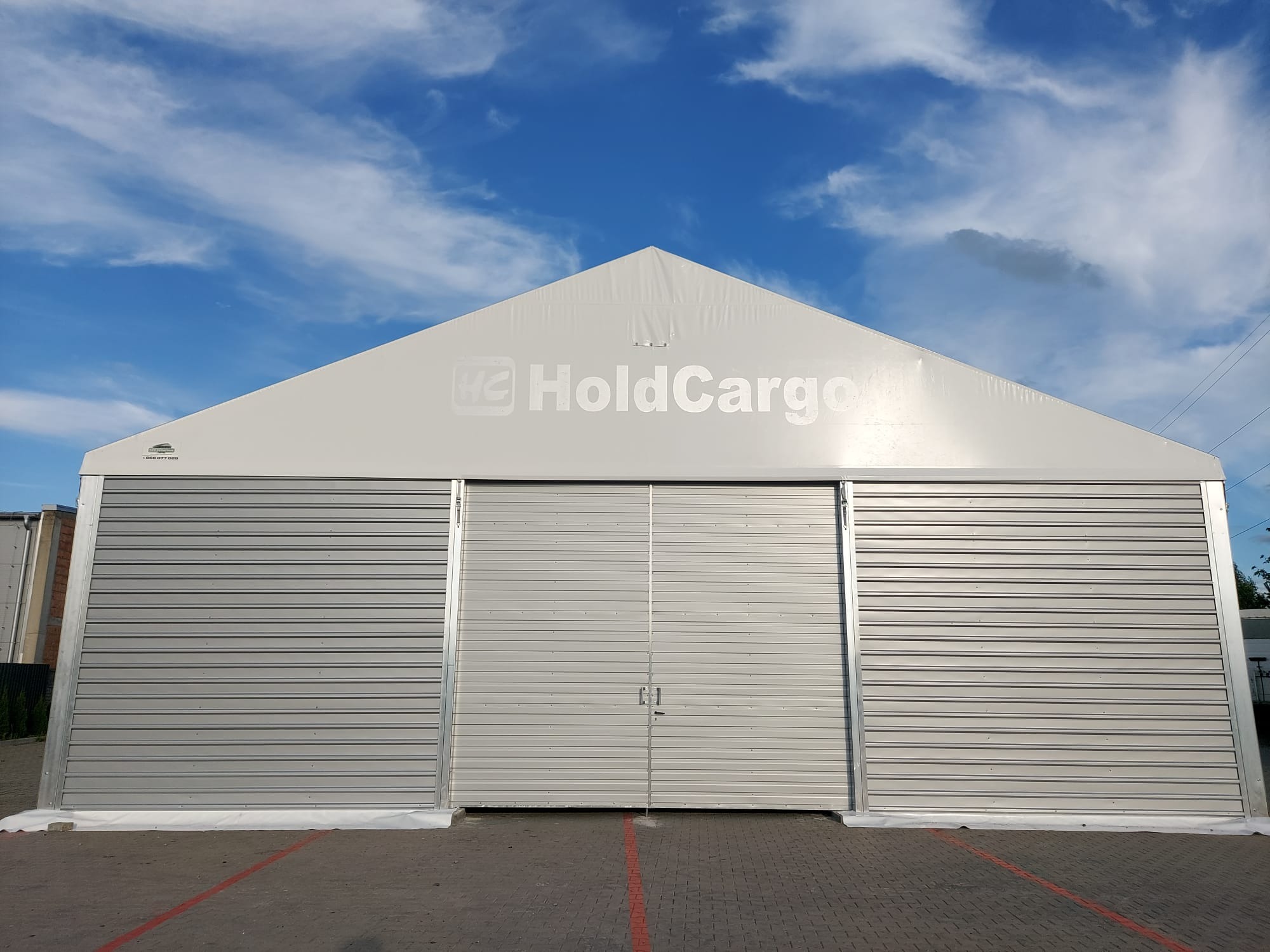Hala magazynowa z blachy trapezowej z logo HoldCargo na tle błękitnego nieba. Widoczne zamknięte wrota i fragment parkingu z liniami. Jasne oświetlenie.