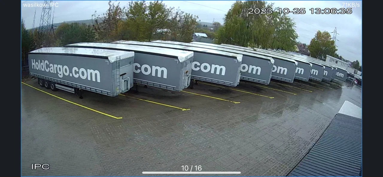 Szereg szarych naczep HoldCargo.com zaparkowanych na mokrym placu, widok z góry pod kątem. Żółte linie wyznaczają miejsca parkingowe. Data i godzina w rogu.