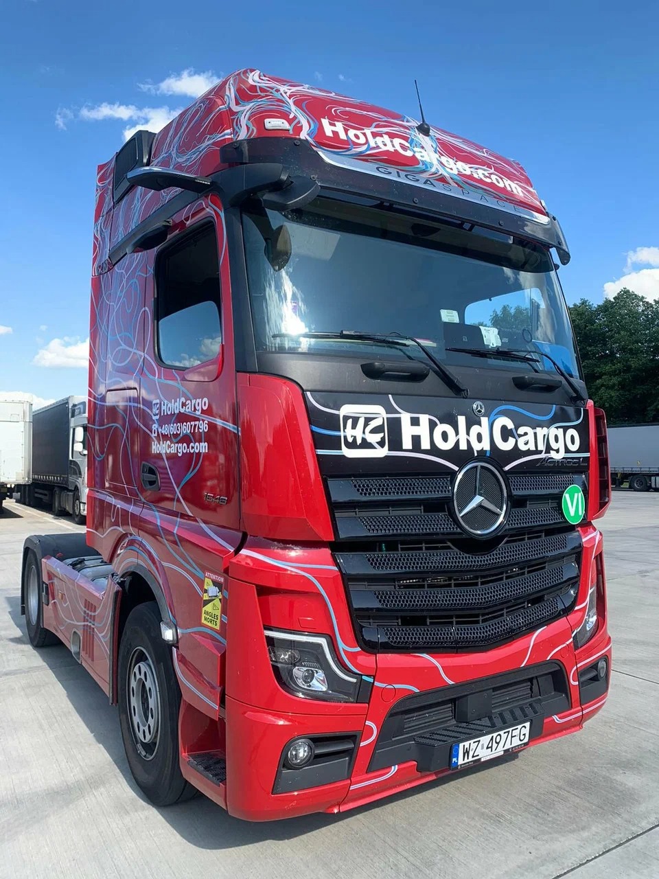 Czerwony ciągnik siodłowy Mercedes-Benz Actros z grafiką Hold Cargo, zaparkowany na betonowym placu w słoneczny dzień. Widoczny numer rejestracyjny WZ 497FG.