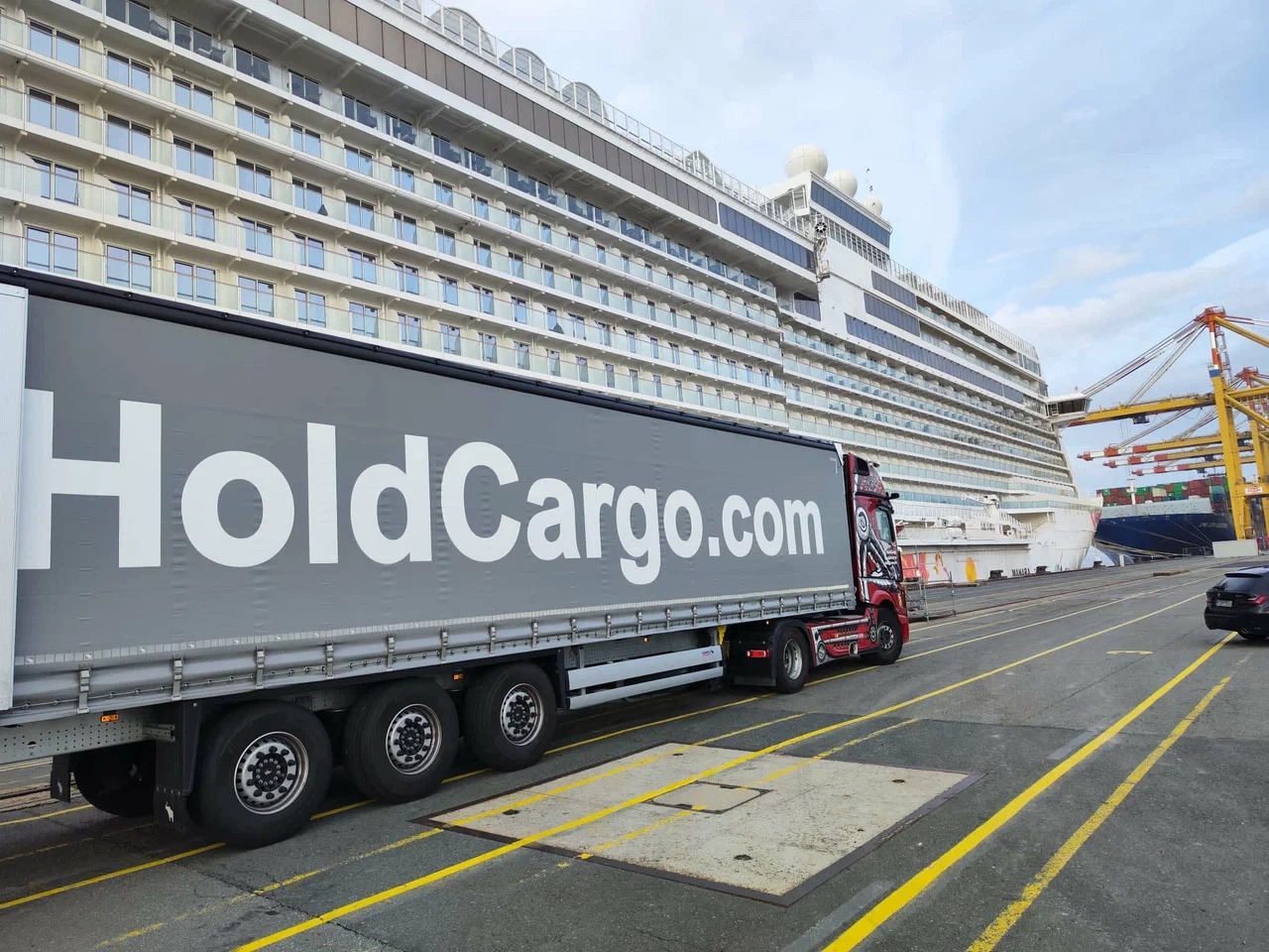 Ciężarówka HoldCargo.com na tle statku wycieczkowego i dźwigów portowych. Szara naczepa, czerwona kabina z grafiką. Perspektywa z dołu, pochmurne niebo.