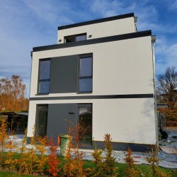 T&O Company - Nowoczesny, trzypiętrowy dom z płaskim dachem, wykończony w bieli i szarości, z czarnymi akcentami. Widoczne młode drzewka na pierwszym planie i pracownik przy elewacji.