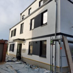 T&O Company - Dwukondygnacyjny budynek z białą elewacją i czarnymi akcentami, widoczne rusztowanie i folia ochronna, sugerujące prace wykończeniowe. Osoba pracuje przy elewacji.