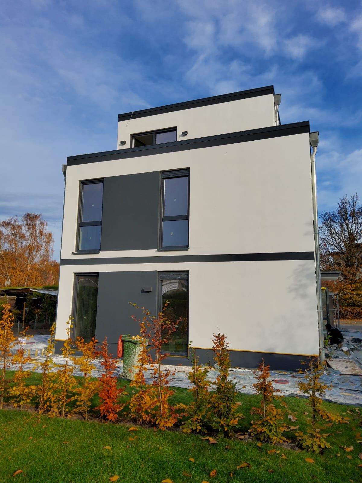 Nowoczesny, trzypiętrowy dom z płaskim dachem, wykończony w bieli i szarości, z czarnymi akcentami. Widoczne młode drzewka na pierwszym planie i pracownik przy elewacji.