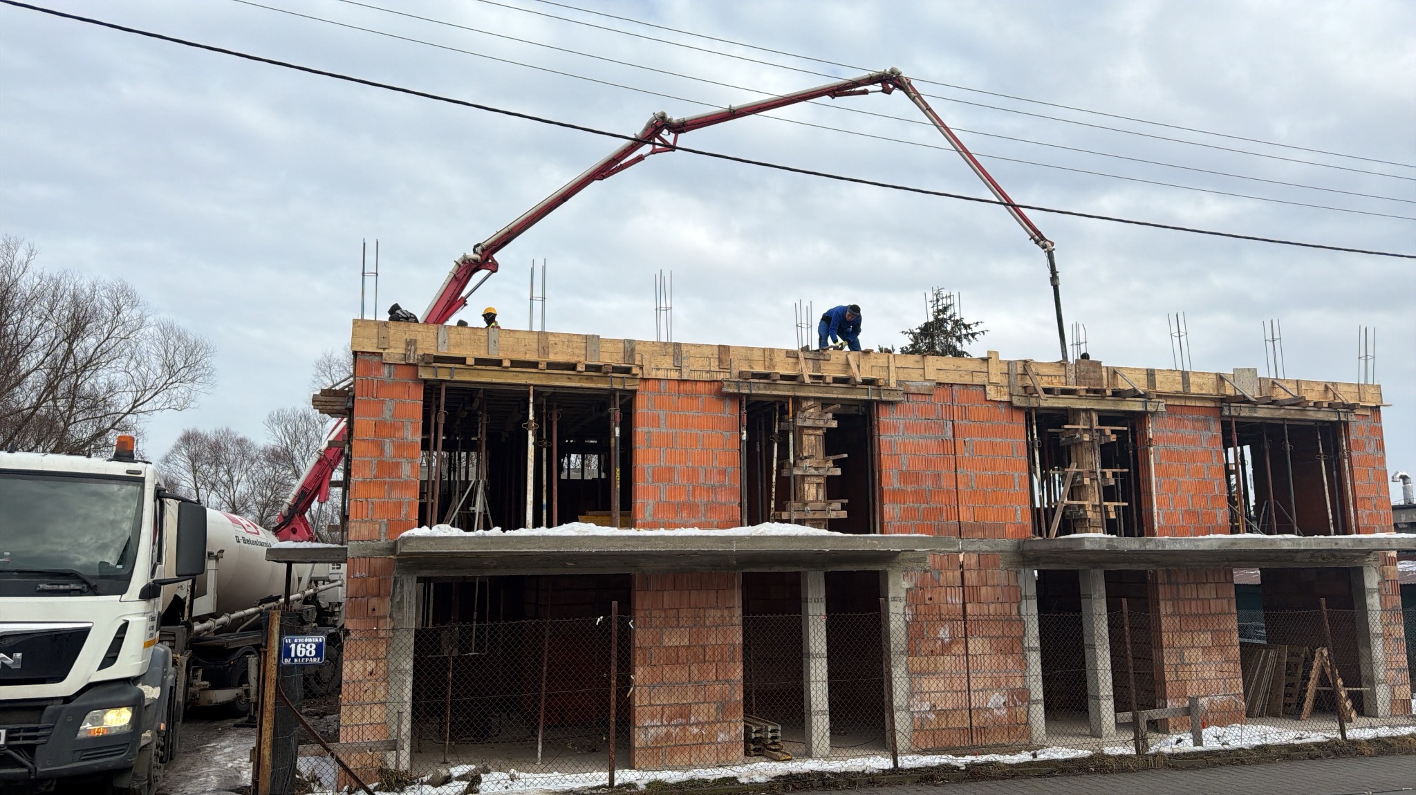 Wylewanie betonu na strop budynku w trakcie budowy. Widoczny żuraw do betonu, pracownicy w kaskach i surowy stan budynku z czerwonej cegły.