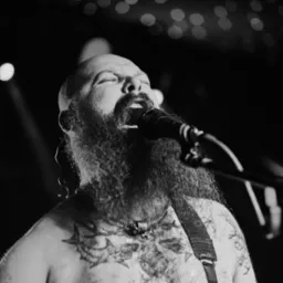 Czarno-białe zdjęcie brodatego mężczyzny z tatuażami, śpiewającego do mikrofonu na scenie z rozmytymi światłami w tle. Ekspresja, emocje, pasja.