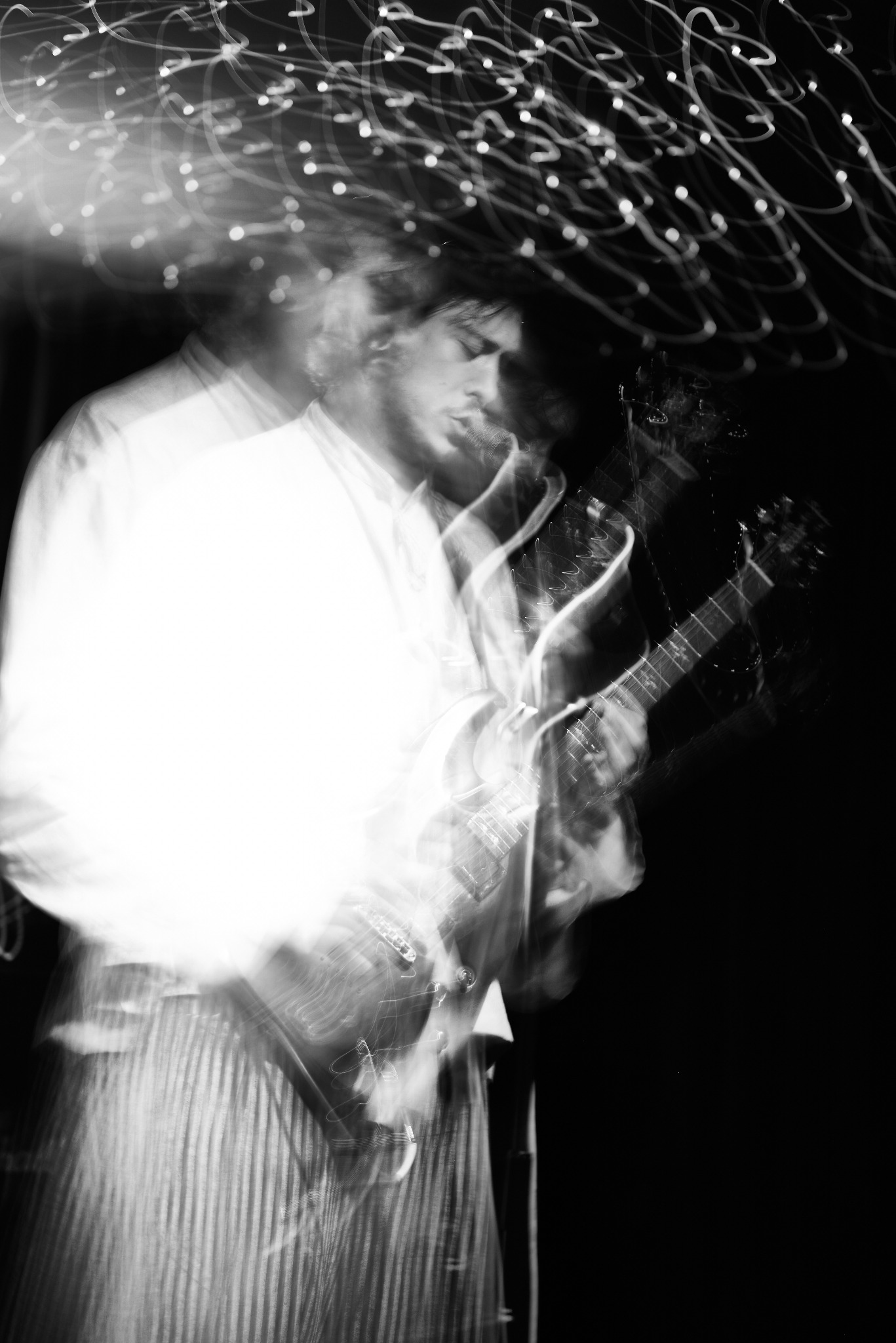 Czarno-białe, rozmyte zdjęcie gitarzysty podczas koncertu. Efektowne smugi świetlne nad głową muzyka dodają dynamiki. Ujęcie z dołu, podkreślające ekspresję.