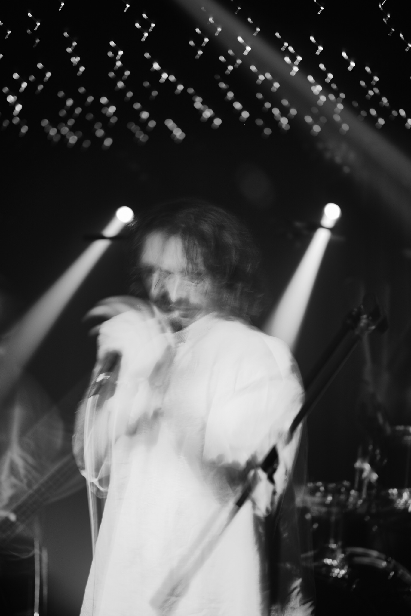 Czarno-białe, rozmyte zdjęcie piosenkarza z mikrofonem na scenie z reflektorami i dekoracyjnymi światłami w tle, uchwycone w ruchu podczas występu.