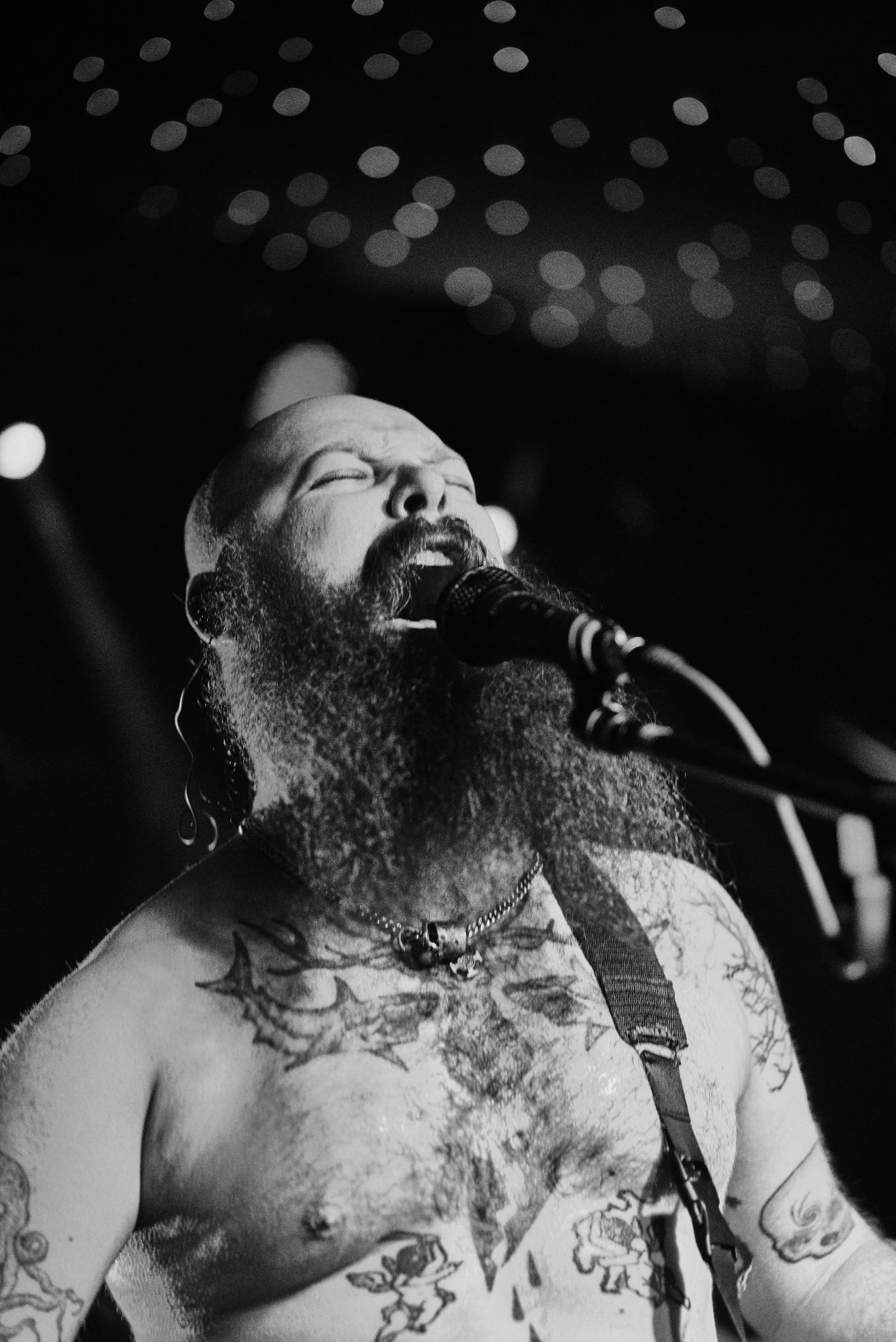 Czarno-białe zdjęcie brodatego mężczyzny z tatuażami, śpiewającego do mikrofonu na scenie z rozmytymi światłami w tle. Ekspresja, emocje, pasja.