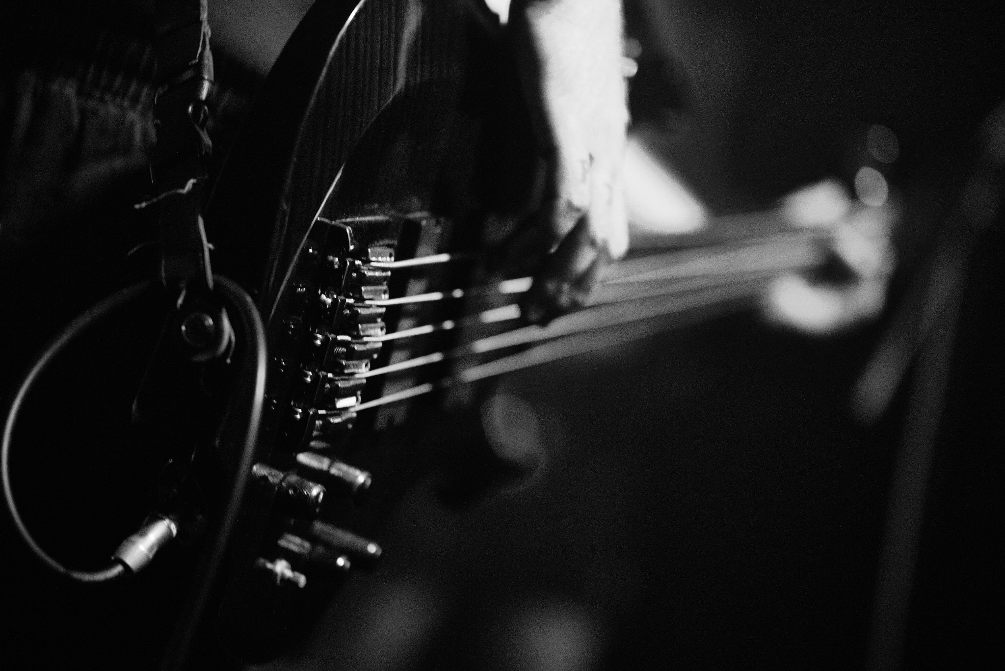 Czarno-białe zdjęcie gitary basowej z widocznymi strunami, mostkiem i słuchawkami, uchwycone w trakcie koncertu, z ręką muzyka w tle.