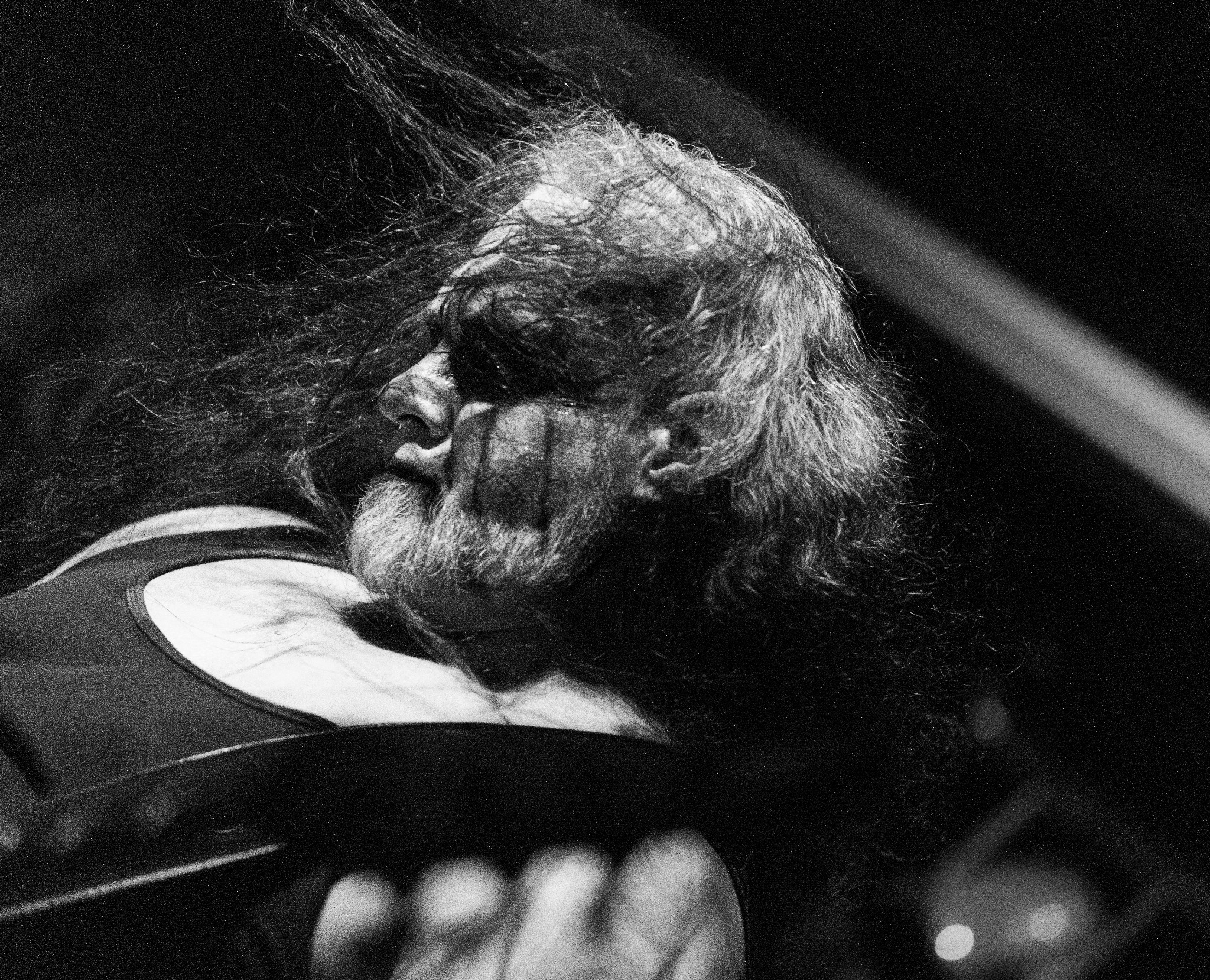 Czarno-białe zdjęcie profilu mężczyzny z długimi włosami i brodą, z ciemnymi plamami na twarzy, prawdopodobnie makijażem, w dynamicznym ruchu, uchwycone w słabym oświetleniu.