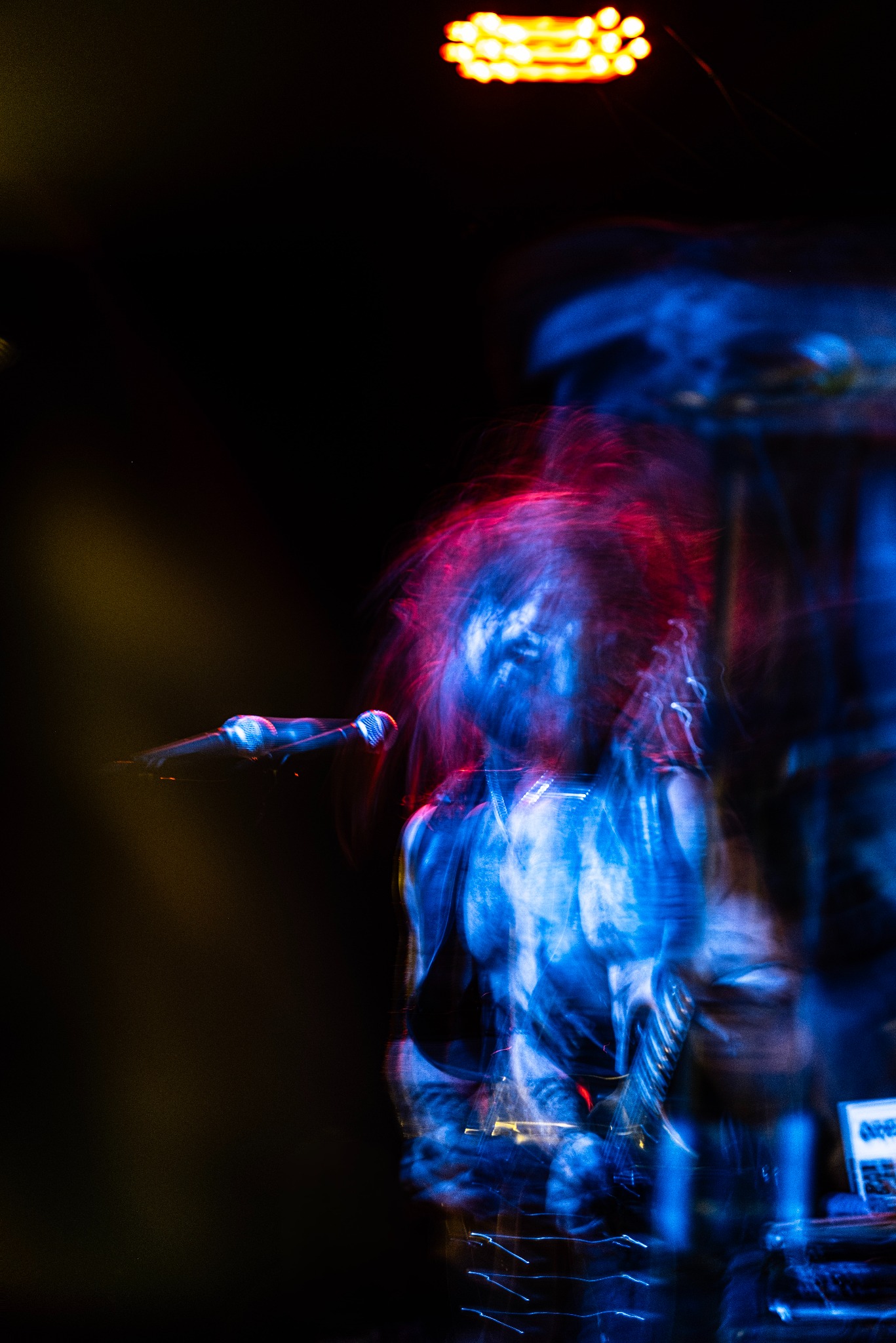 Dynamiczne, rozmazane ujęcie piosenkarza z mikrofonem na scenie w intensywnym, niebiesko-czerwonym oświetleniu eventowym. Widoczny fragment sprzętu muzycznego.