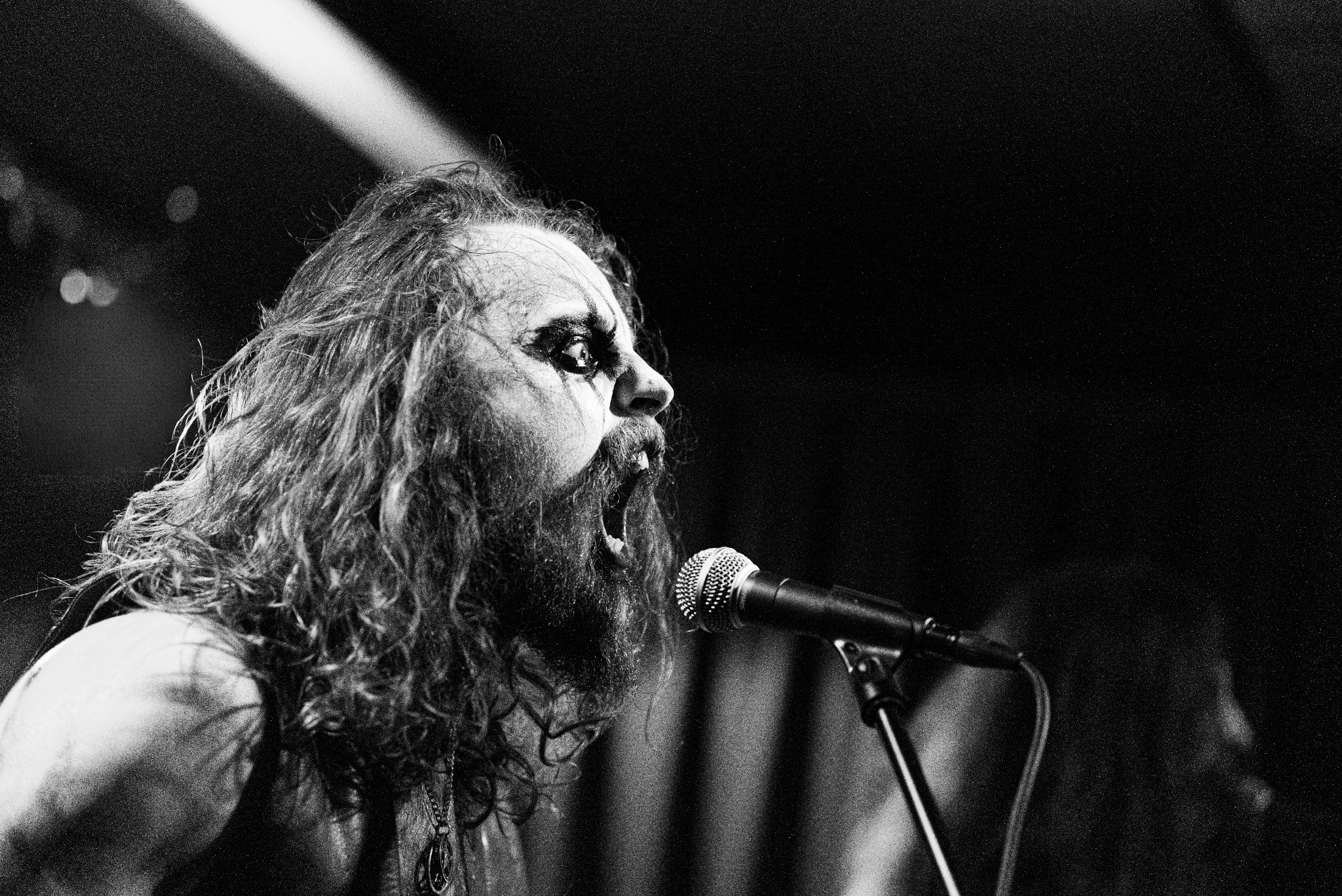 Czarno-białe zdjęcie profilu wokalisty z długimi włosami i makijażem scenicznym, śpiewającego do mikrofonu podczas występu na żywo w ciemnym otoczeniu.