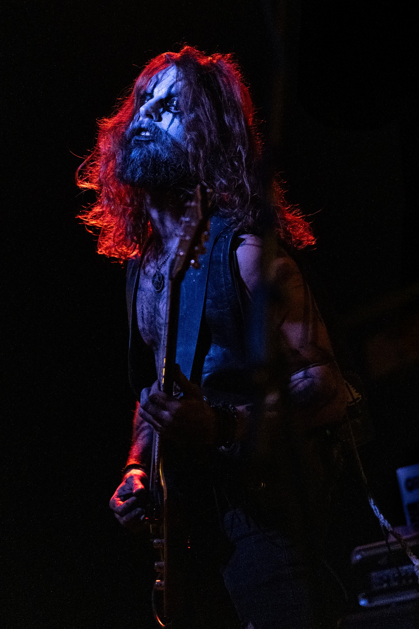 Mężczyzna z długimi włosami i makijażem scenicznym gra na gitarze elektrycznej podczas koncertu w ciemnym otoczeniu, oświetlony czerwono-niebieskim światłem.