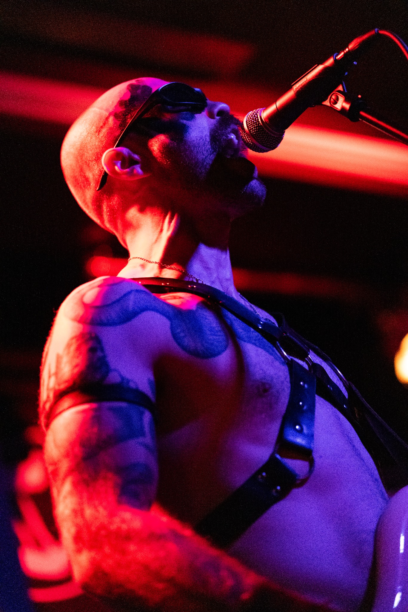 Wokalista z tatuażami śpiewa do mikrofonu w czerwono-fioletowym oświetleniu scenicznym. Ubrany w skórzane szelki, z okularami przeciwsłonecznymi na głowie.