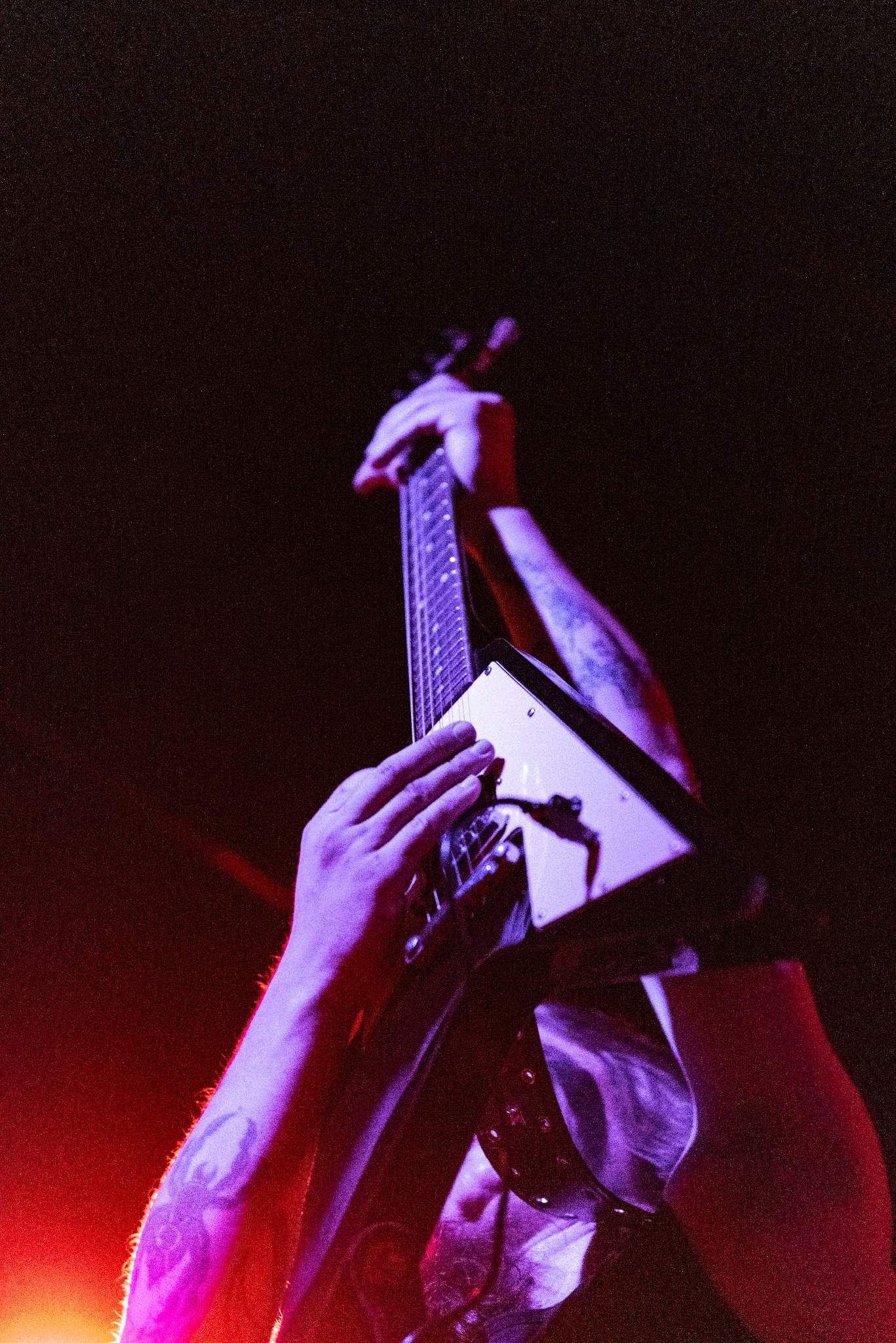 Gitarzysta z tatuażami gra na białej gitarze typu V w czerwono-fioletowym oświetleniu scenicznym. Ujęcie z dołu, podkreślające ekspresję artysty.