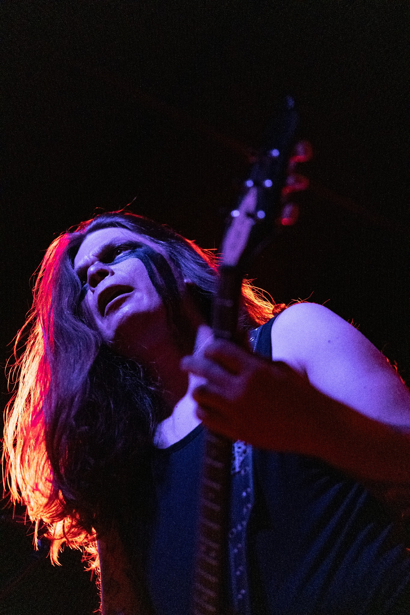 Wokalista black metalowy z gitarą na scenie, z charakterystycznym makijażem oczu, oświetlony czerwono-fioletowym światłem podczas występu.