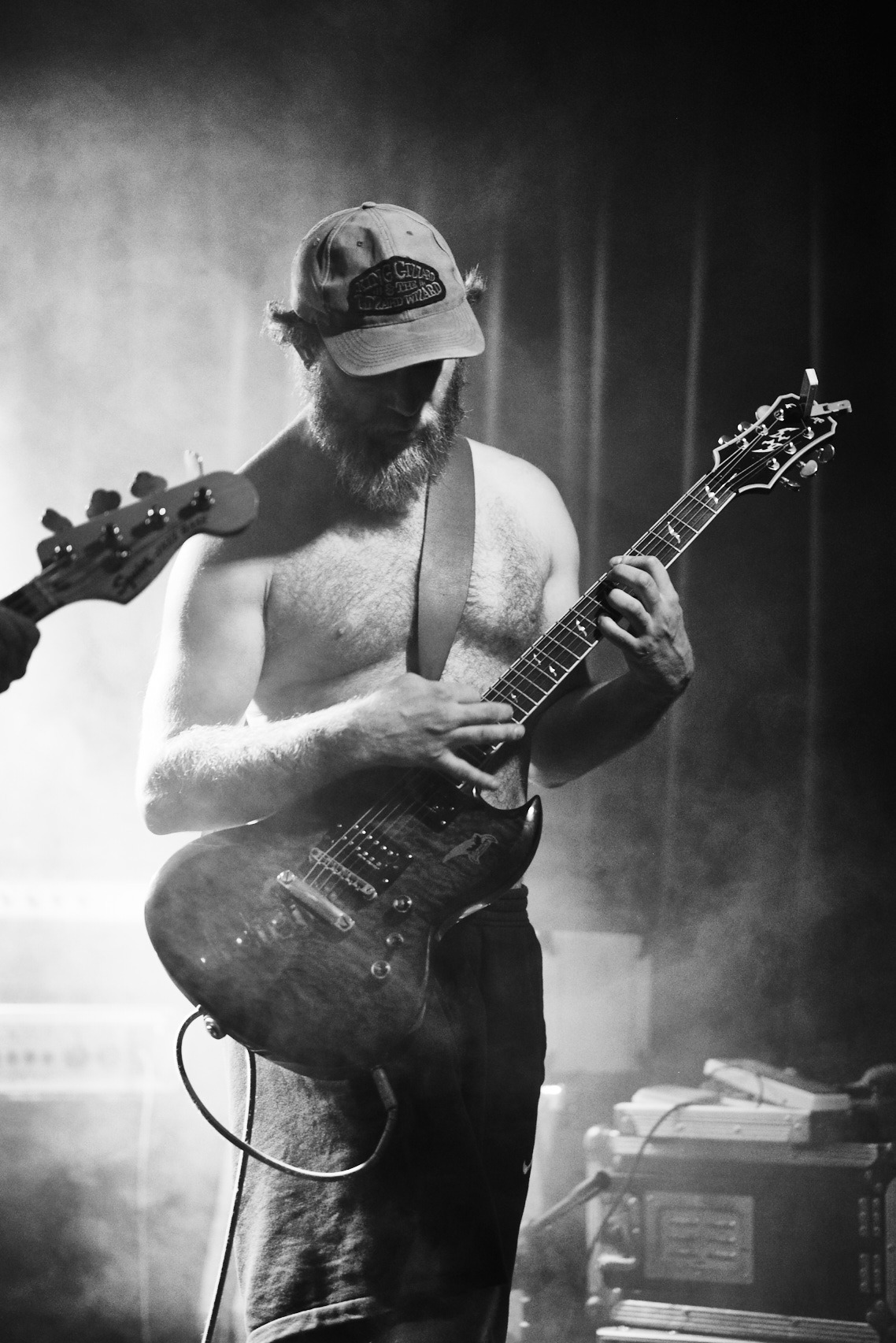 Czarno-białe zdjęcie brodatego gitarzysty bez koszuli, w czapce, grającego na elektrycznej gitarze podczas koncertu. W tle widoczny basista i sprzęt nagłośnieniowy.