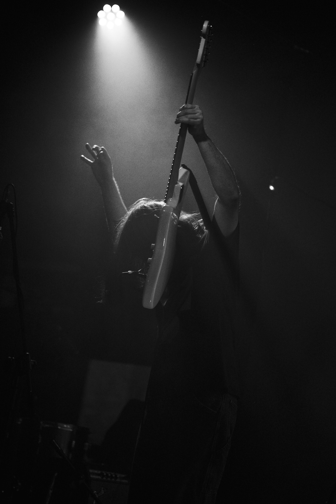 Czarno-białe zdjęcie gitarzysty z podniesioną gitarą podczas występu na scenie, oświetlonego punktowym światłem.