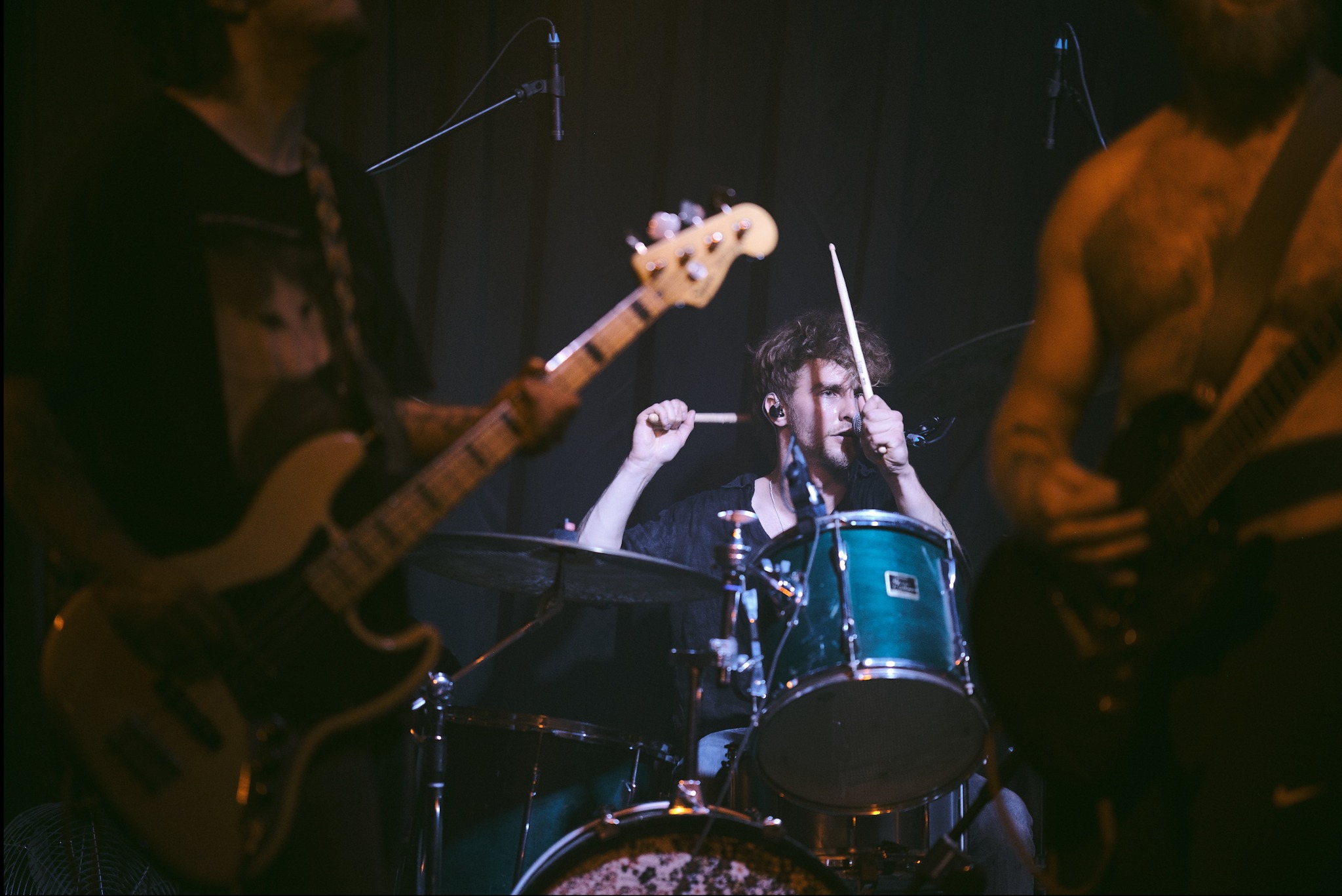 Zespół muzyczny podczas występu na scenie: perkusista gra na bębnach, gitarzysta basowy i gitarzysta elektryczny po bokach. Scena oświetlona punktowo.