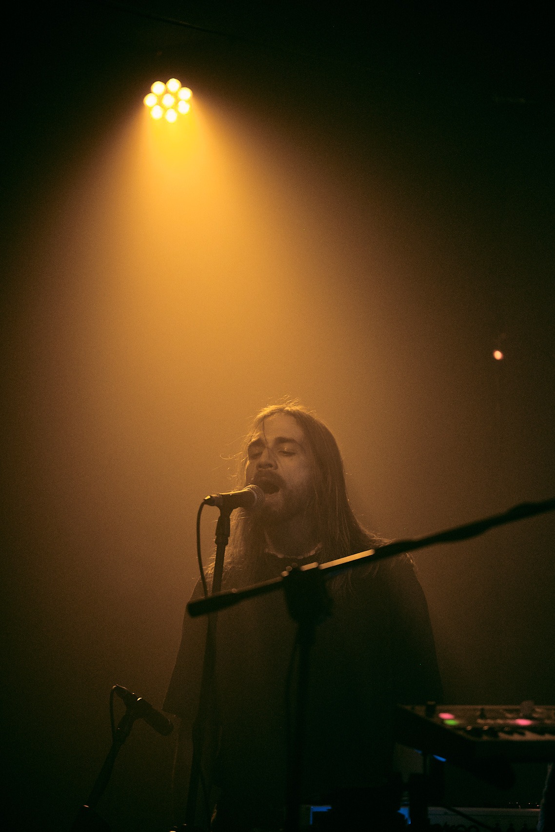 Wokalista z długimi włosami śpiewa do mikrofonu w pomarańczowym świetle reflektorów na scenie. Widoczny fragment instrumentu klawiszowego.