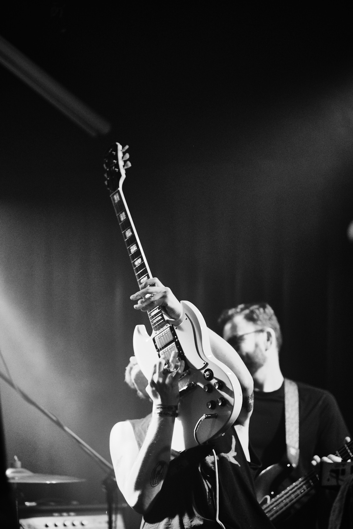 Czarno-białe zdjęcie muzyka z gitarą elektryczną na scenie podczas koncertu, z drugim muzykiem w tle, oświetlonym punktowo.