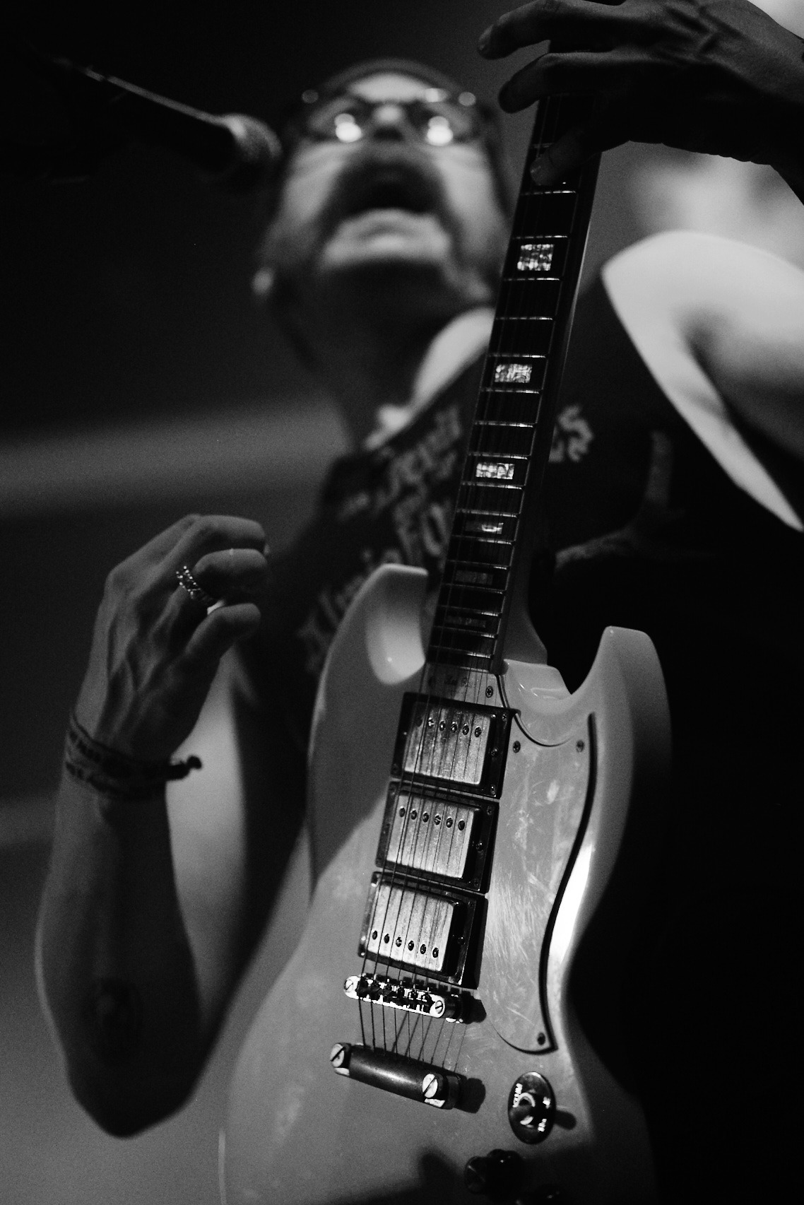 Czarno-białe ujęcie gitarzysty z wąsami, śpiewającego do mikrofonu, z gitarą elektryczną na pierwszym planie. Wyraziste oświetlenie sceniczne.