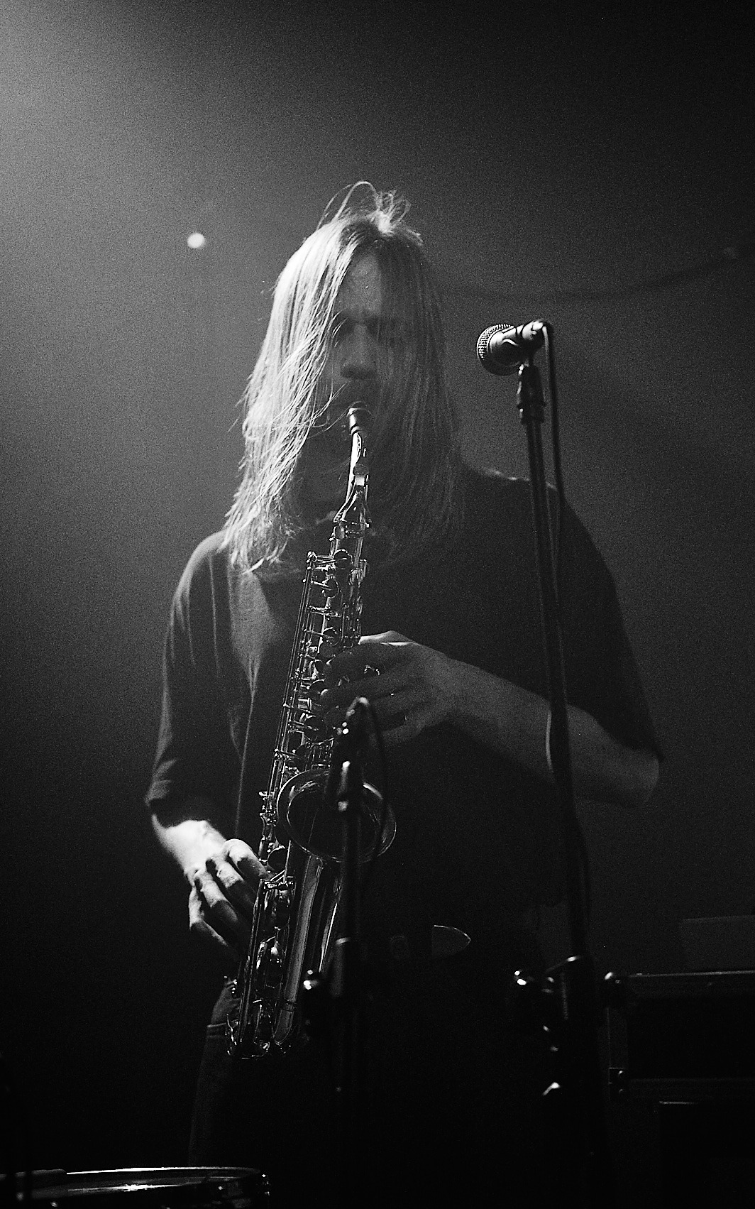 Czarno-białe ujęcie saksofonisty z długimi włosami podczas występu na scenie, z mikrofonem w tle. Oświetlenie sceniczne tworzy dramatyczny efekt.
