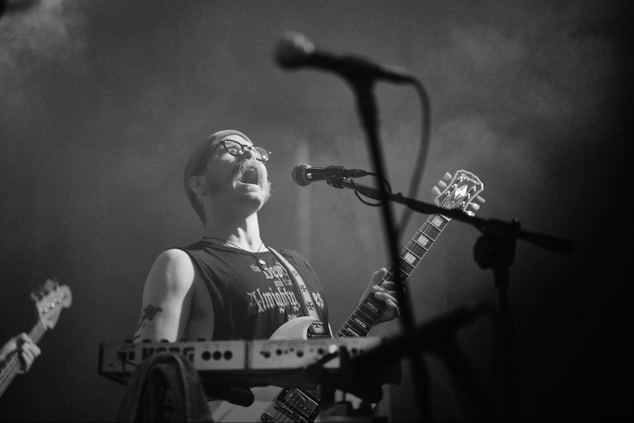 Czarno-białe zdjęcie muzyka z wąsami w beanie, grającego na gitarze elektrycznej i śpiewającego do mikrofonu podczas koncertu na scenie.